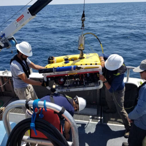 Four people on a boat handle a yellow autonomous underwater vehicle attached to a crane over open lake water.